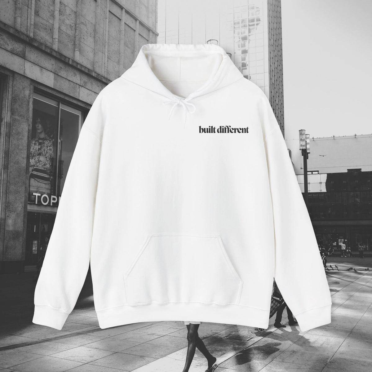 White Built Different unisex pullover hoodie with black chest lettering, photographed against a city street background