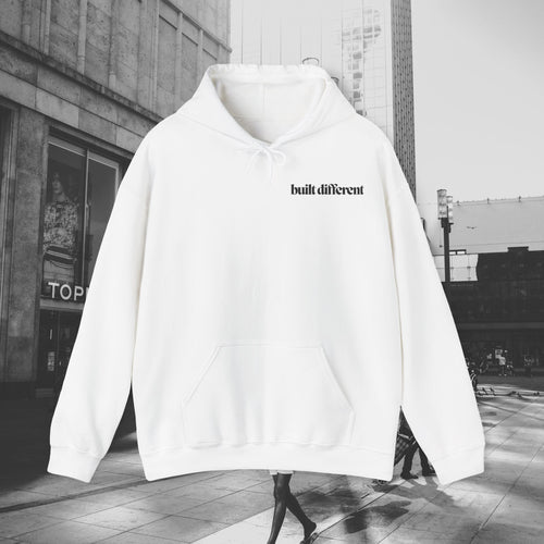 White Built Different unisex pullover hoodie with black chest lettering, photographed against a city street background